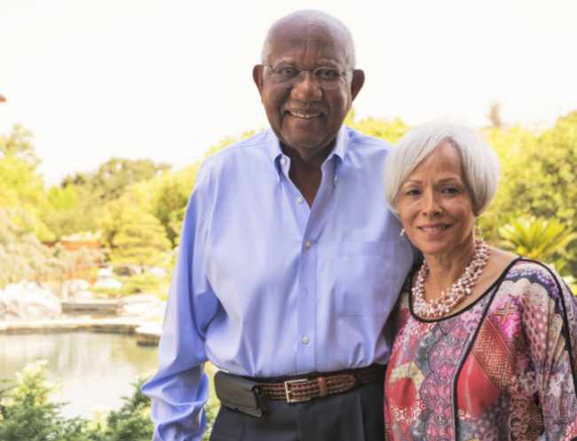 An older Black gentleman stands in a light blue dress shirt aside his wife, who is dressed in a multi-colored blouse. They are arm in arm and standing outdoors in front of a pond and greenery.