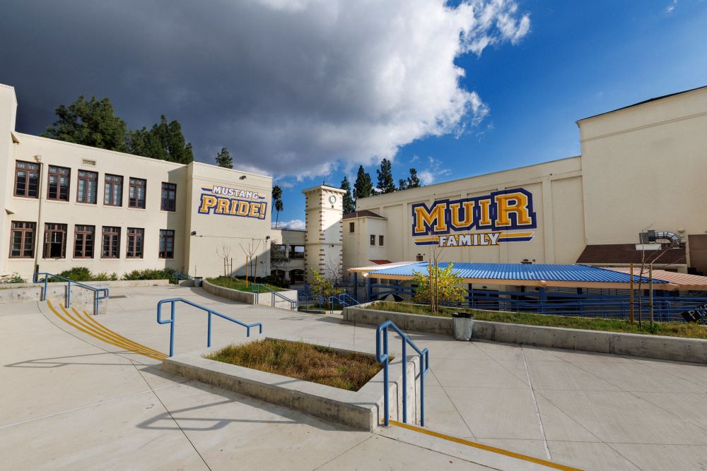 Two high school buildings and a plaza are shown with a dramatic sky and clouds above them. One building has "Muir Family" painted in blue and yellow and the other building has "Mustang Pride" painted in blue and yellow. 