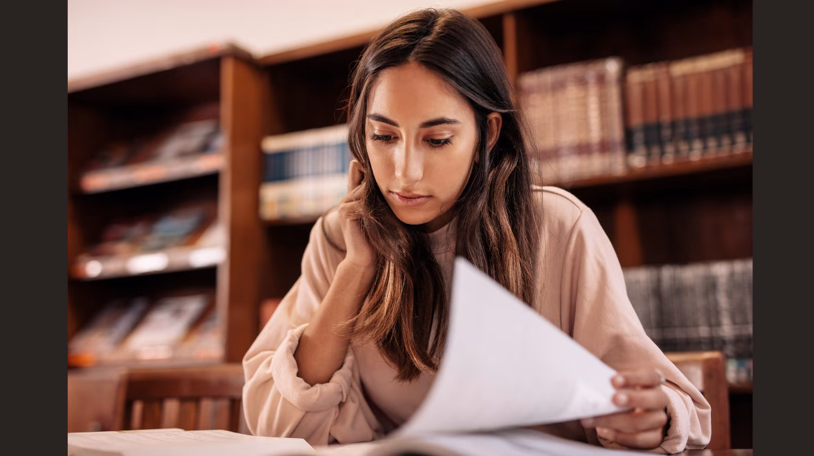 A college age woman sits in a library studying a paper at a table