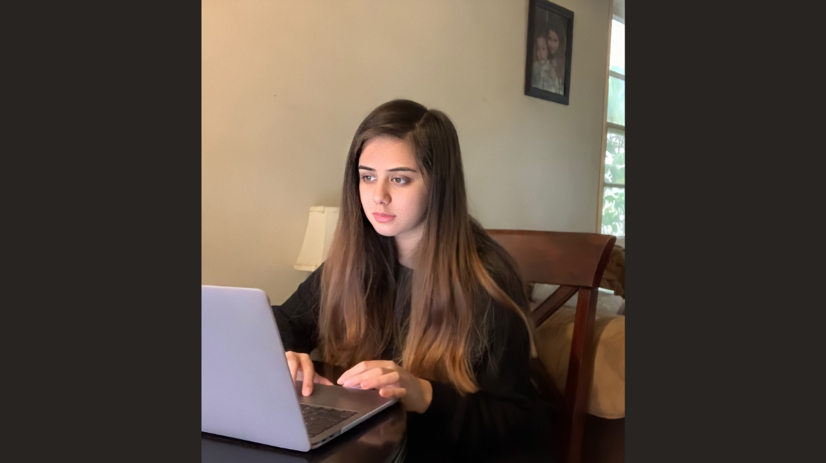 A young woman with long dark hair sits inside a room looking at a laptop