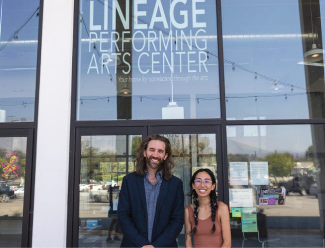 A man and a woman stand outside glass doors and windows with a sign that reads Lineage Performing Arts Center.