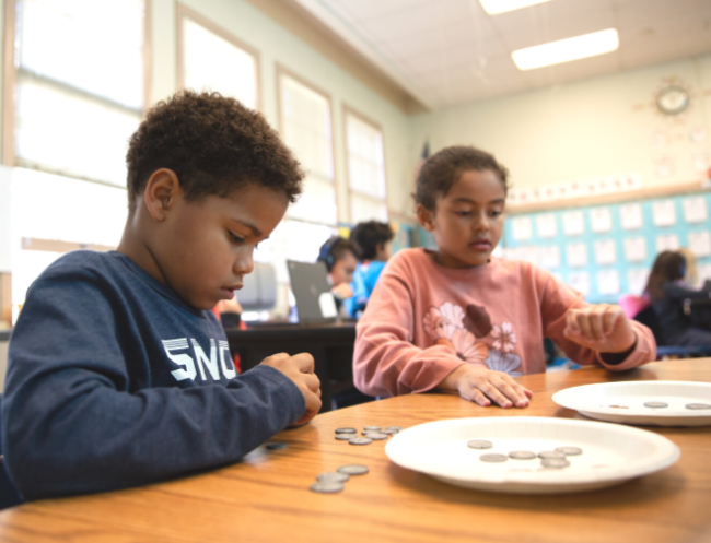 Two Black children sit at a schoolroom table counting out coins as part of a math lesson