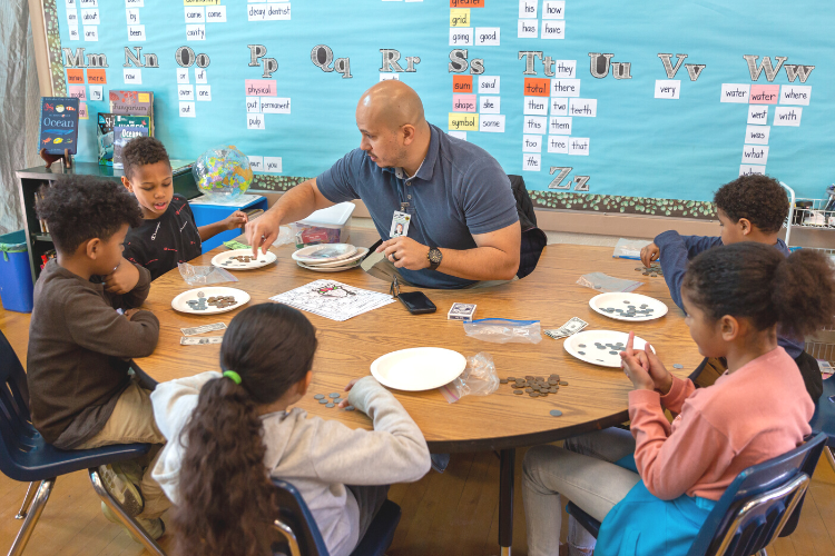 An adult volunteer sits at a circular table inside a Pasadena second grade classroom. He has five students gathered around him at the table, each working on math concepts using coins and dollar bills. 