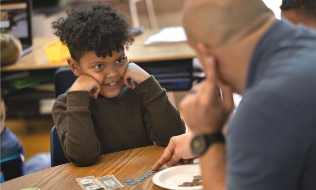 A young Black student sits at a table, hands up supporting his chin, looking eagerly up to a male adult, who is helping him count out change and currency.