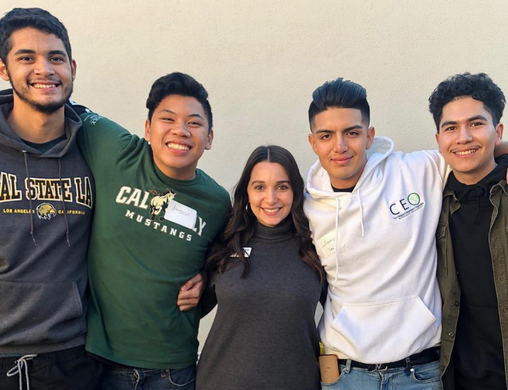 Four male high school students wearing college-themed sweatshirts stand embracing together in a line, smiling and facing the camera. A woman with a gray sweatshirt stands in the middle of them.