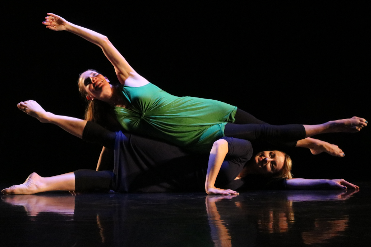 Two women dancers in leotards perform while nearly lying horizontal on a darkened stage.