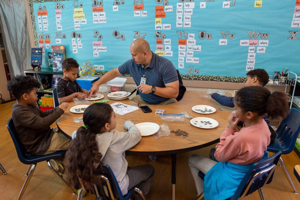 A male teacher sits at a round table with several young elementary school aged children. A colorful blue bulletin board is behind him