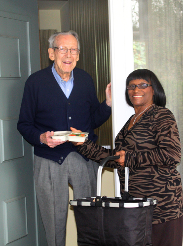 A woman stands at a front door holding a basket of food. She is delivering Meals on Wheels to an elderly gentleman client. 