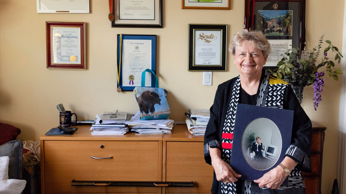 An elderly woman wearing a black, gray and red dress, stands inside a home office holding a photo of herself in earlier years 