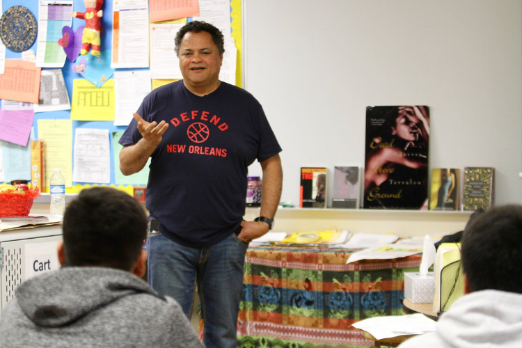 An older Latino man wearing a "Defend New Orleans" tshirt stands in front of a classroom. His hand is gesturing, and he appears to be engaged in a conversation with a student in the front row.