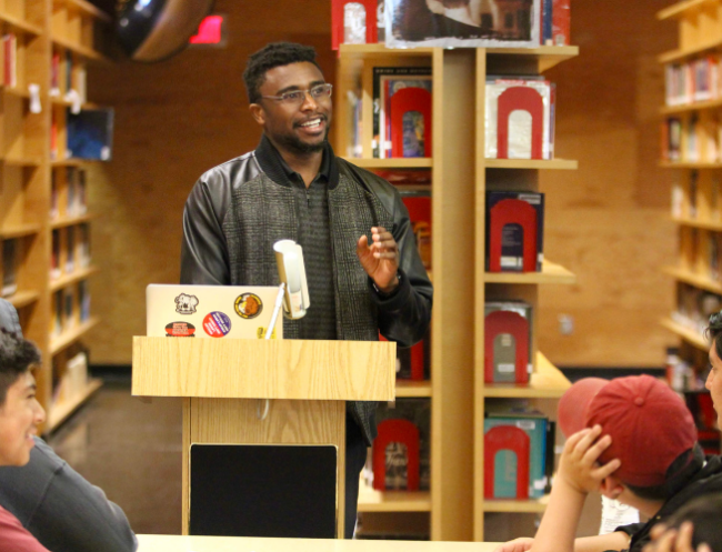 A young Black man in glasses stands at a podium inside a library. Bookshelves are visible behind him. He is addressing a group of younger students.