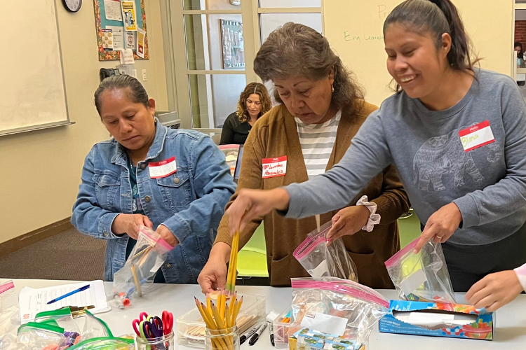 Three Latina women stand at a table inside a classroom. They are reaching for jars of pencils and scissors and filling Ziploc bags. Each wear a red and white nametag.  A woman is visible behind them, seated at the classroom table. 