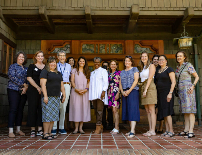 A group of 13 people stand in pose on the front porch of the historic Gamble House in Pasadena CA. They are grouped loosely in a line and surrounded by wooden craftsman architecture.