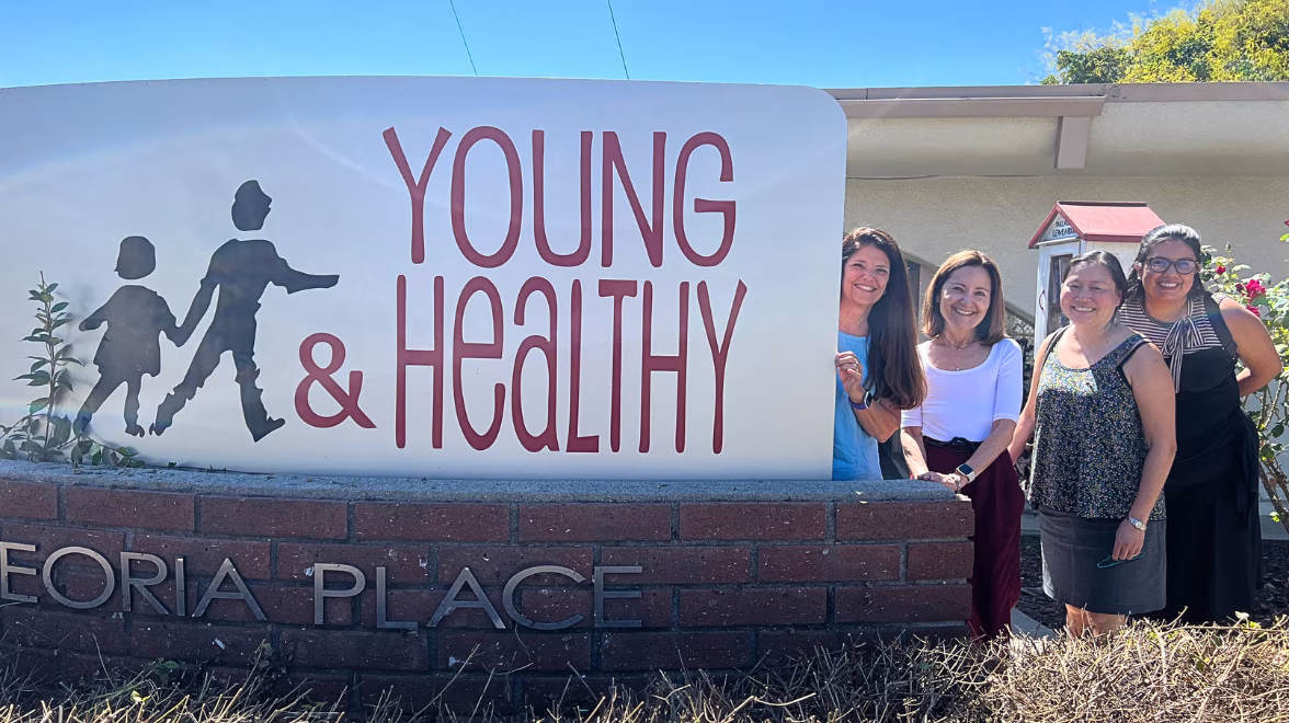 four women stand outdoors partially behind a large sign that reads Young & Healthy