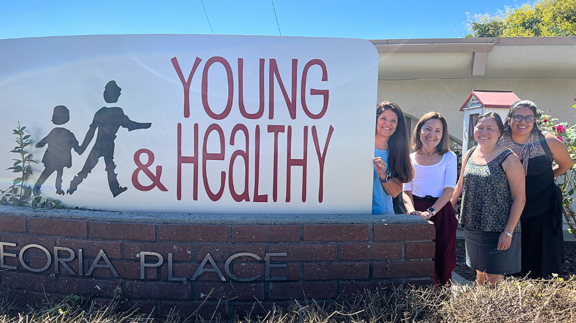 four women stand outdoors partially behind a large sign that reads Young & Healthy