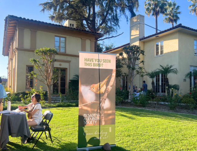 A laughing woman sits at a check-in table to the far left of the photo. Behind her are two two-story buildings with a tall vertical sign on a green lawn that reads “Have you seen this bird?” and shows a photo of a bird in a tree.
