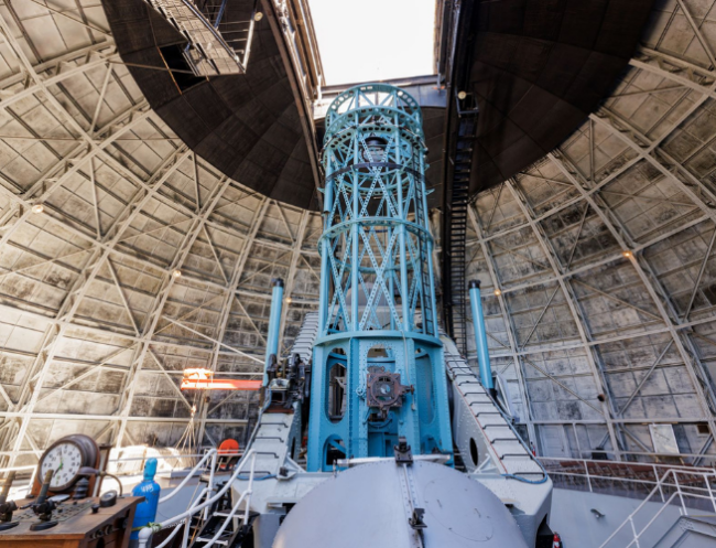 A large telescope painted bright blue stands in the middle of a domed observatory.