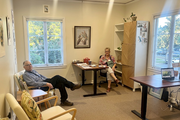 Two adults sit relaxed and comfortably in an office with three chairs, a desk, and tall shelving. They are smiling and looking at the camera. Two windows show that it is daylight outside. 