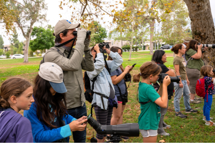 A man holding binoculars to his eyes faces the right side of the photo. Around him are schoolchildren, many of whom are also using scopes and binoculars to search for birds. They are outdoors under a tree in a park-like setting. 