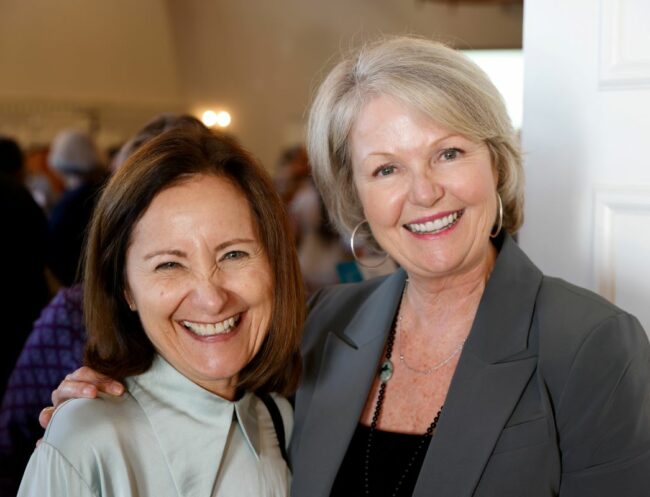 Two women stand together smiling broadly. Their faces and shoulders take up most of the frame. The woman on the right has her arm draped on the shoulders of the woman on the left. They appear to be in a large event room with other guests behind them.