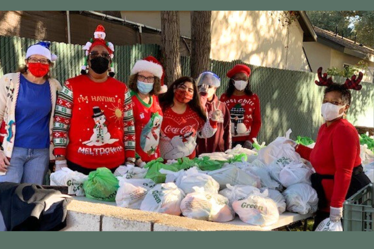 Five women wearing bright, festive holiday attired, including hats and face masks, stand outdoors and to the left of a large pile of bags ready to donate during a food drive. To the right of the bags stands another woman, wearing a red sweater, reindeer antlers, and a white face mask. 