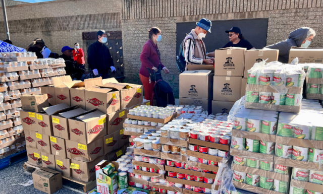 A line of people forms behind large crates and platforms of donated canned goods for a holiday food distribution. They are outdoors, standing in the sunshine.