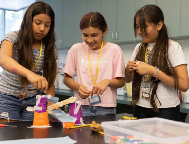 Three girls around the age of 12-13 stand closely together around a table smiling and giggling as they work together to assemble a project.