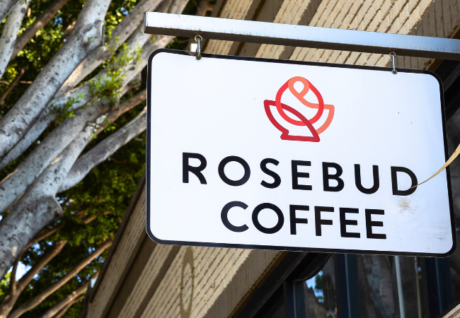 A black and white sign with a stylized rose reads Rosebud Coffee. It appears to be hung outdoors in a tree.