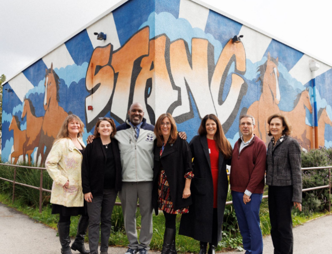 A group of 7 adults stand together facing the camera in front of the corner of a building that painted in a blue, yellow and orange mural that reads MUSTANGS.