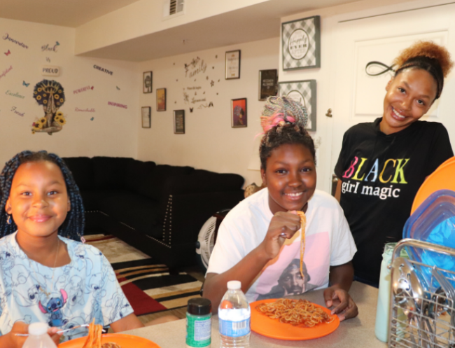A mother stands casually at the far right, leaning on a table and smiling at the camera. Her two daughters sit to the left, eating a meal and smiling at the camera.