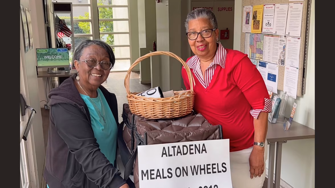Two older Black women stand on either side of a cart with a basket on top. A sign reads Altadena Meals on Wheels.