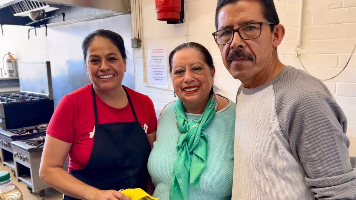 Three Latino adults stand inside a communal kitchen