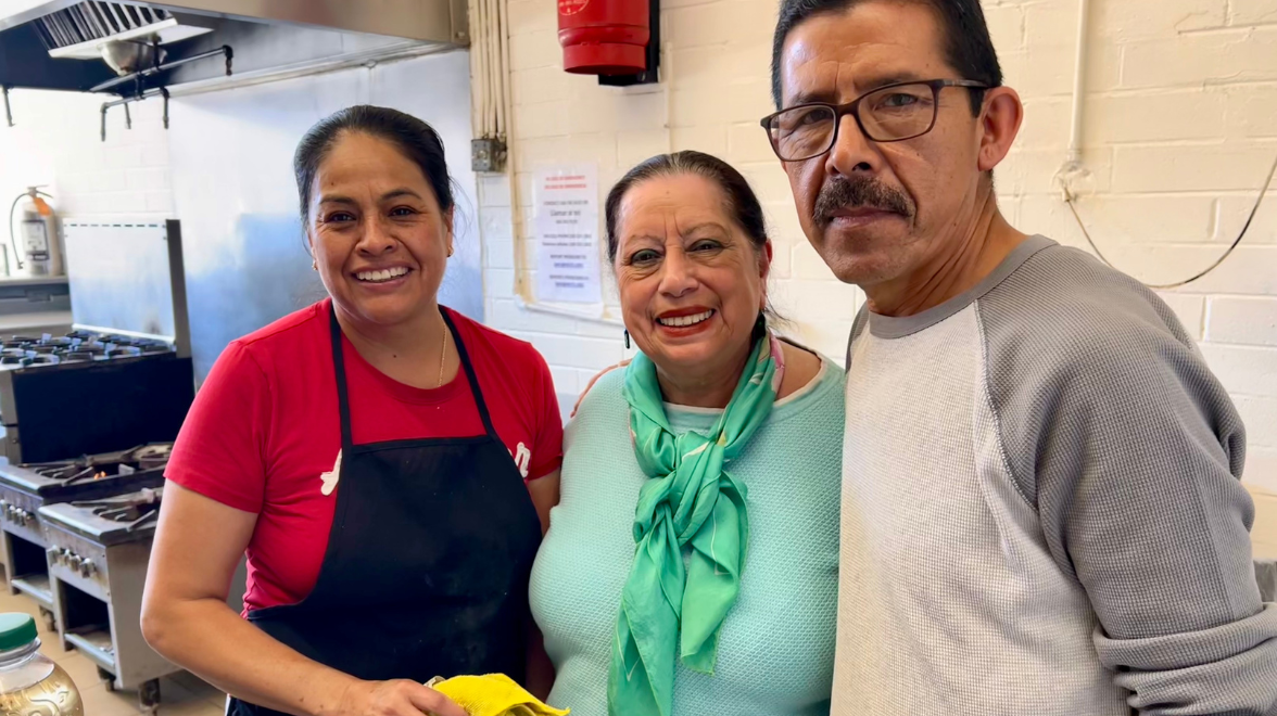 Three Latino adults stand inside a communal kitchen