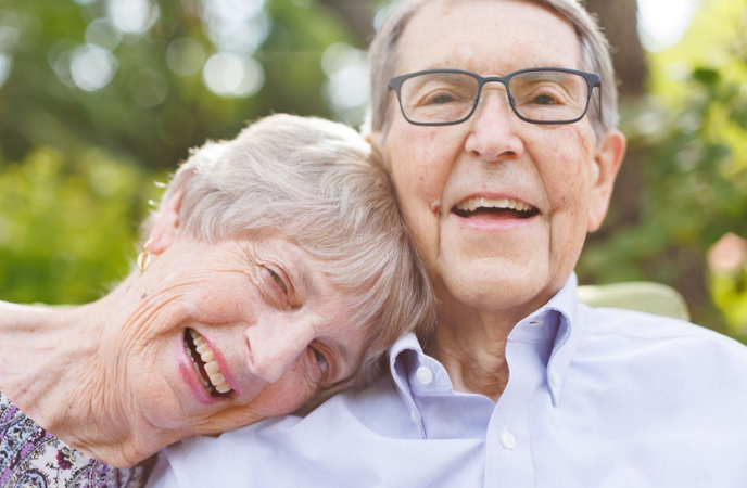 In a close-up photo of their faces, an elderly white couple sits closely, the woman has her head on the shoulder of the man and they are smiling comfortably