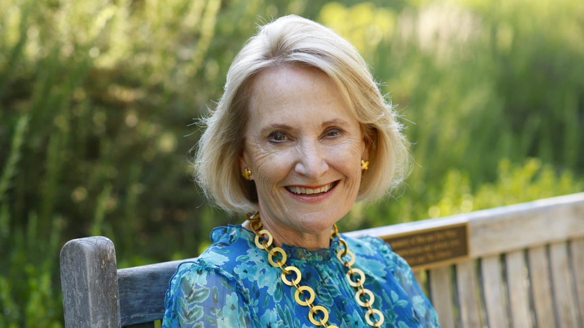 An older white woman in a bright blue dress and chunky gold necklace sits on a park bench