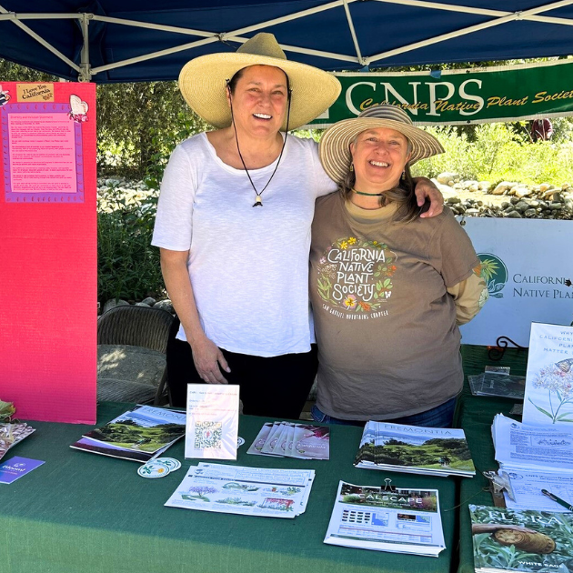 two women stand arm in arm inside a plant event tent