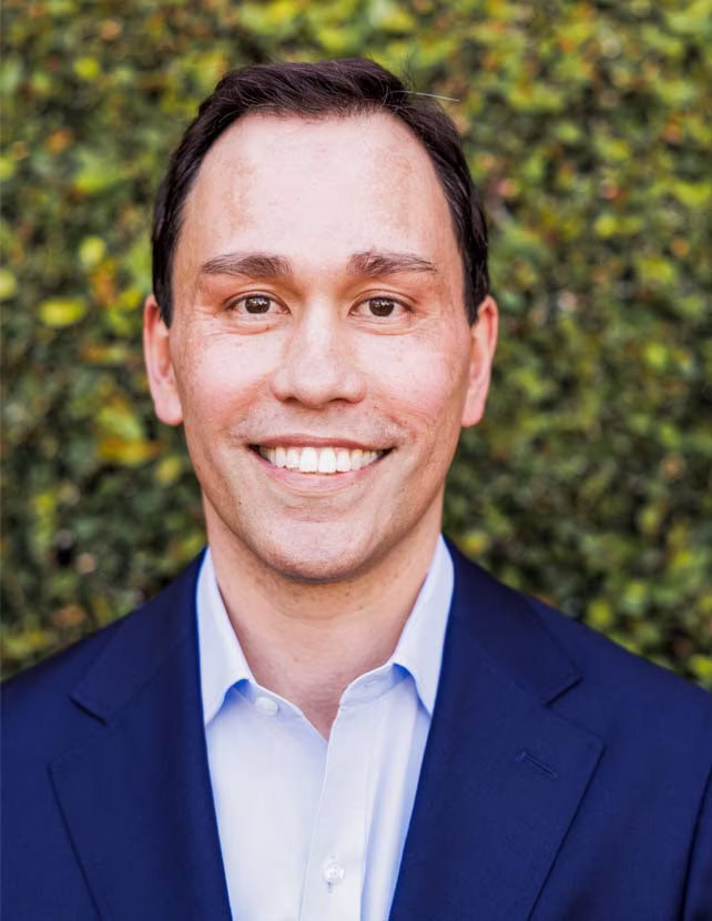 A man in a blue suit is smiling. Green leaf background.