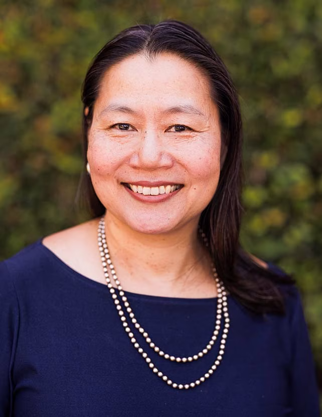 A woman in a necklace and blue shirt is smiling, Green leaf background.
