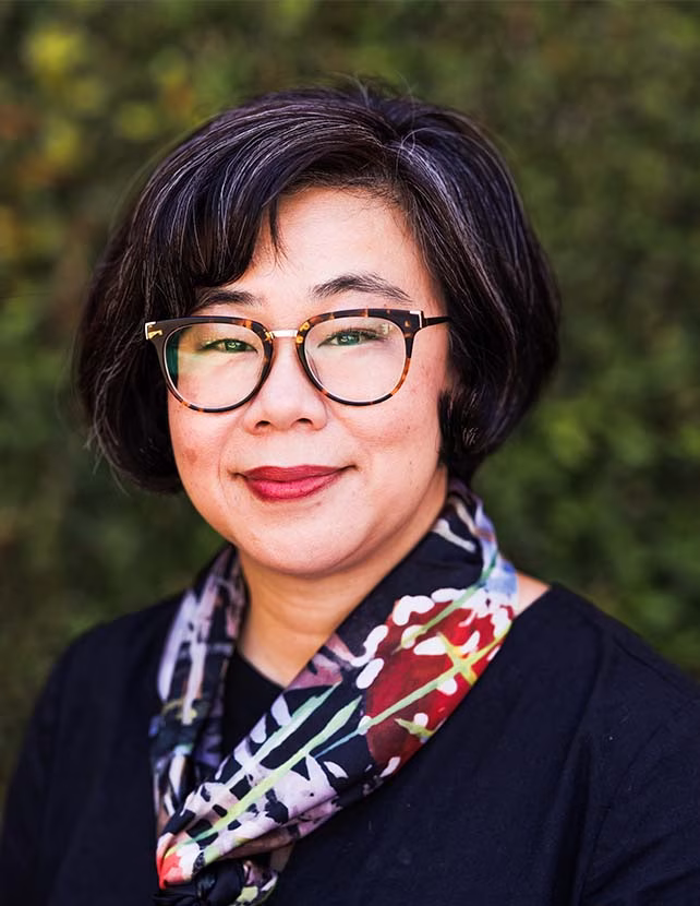 A woman is smiling wearing a black shirt and multi-color scarf. She has on red lipstick. Green leaf background.