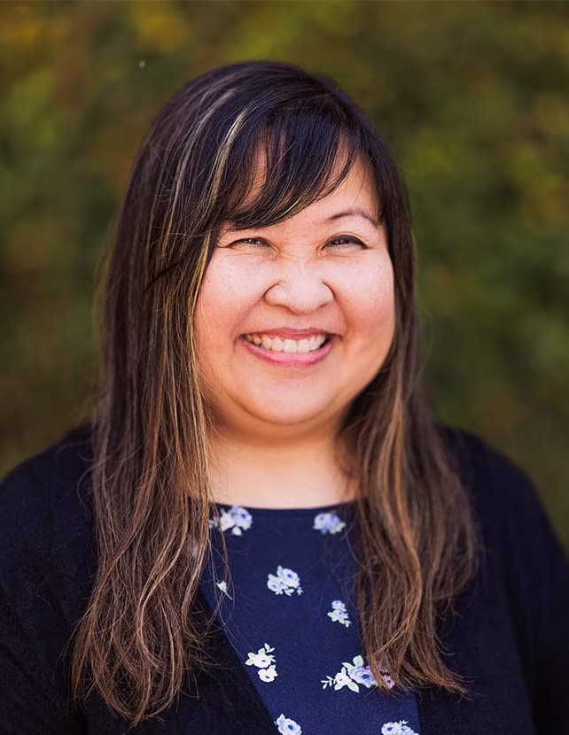 A woman in a black sweater and floral blue top is smiling, Green leaf background.