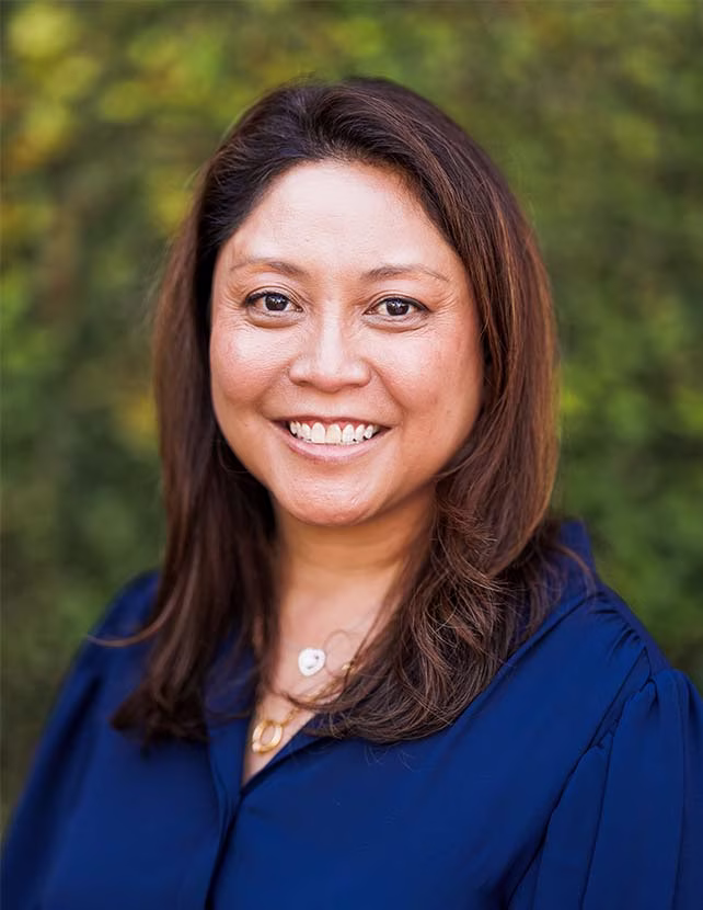 A woman in a dark blue button up shirt is smiling. Green leaf background.