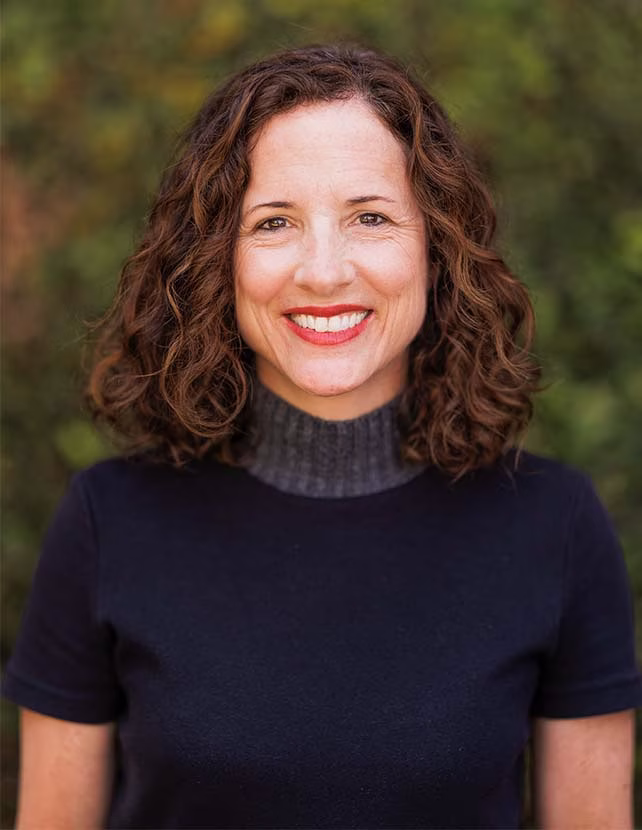 A woman in a black shirt is smiling. Green leaf background.