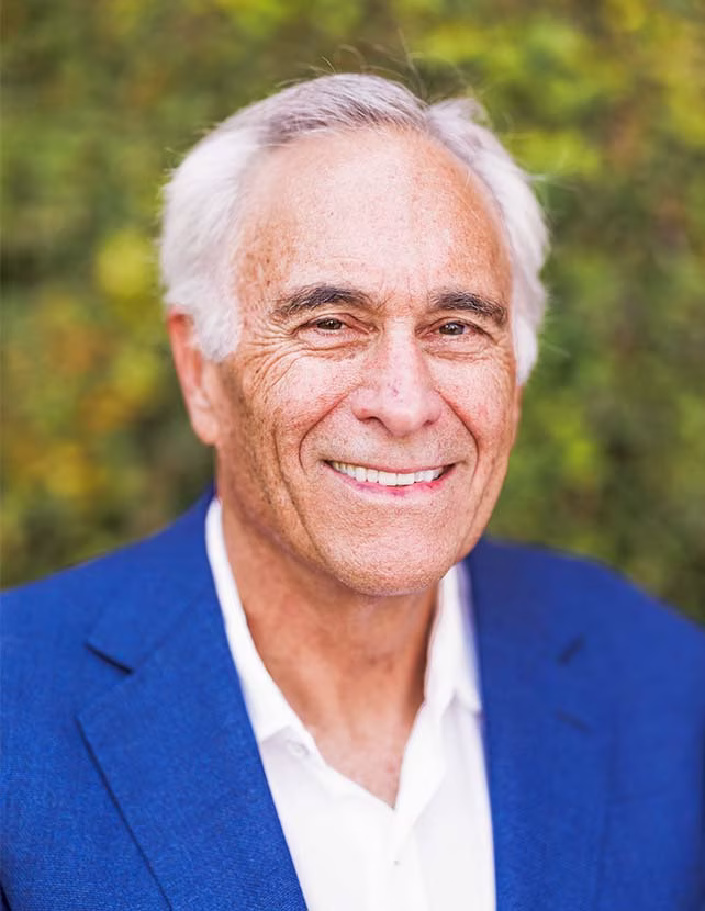 A man is smiling in a blue blazer. Green leaf background.