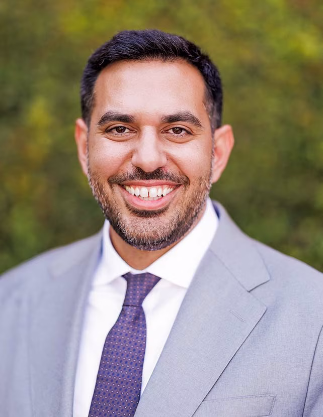 A man in a grey suit is smiling. Green leaf background.
