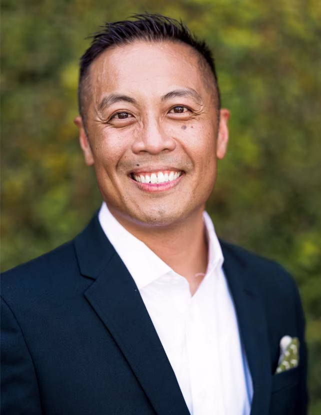 A man in a suit is smiling at the camera. Green leaf background.