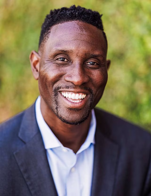 A man in a black suit is smiling. Green leaf background.