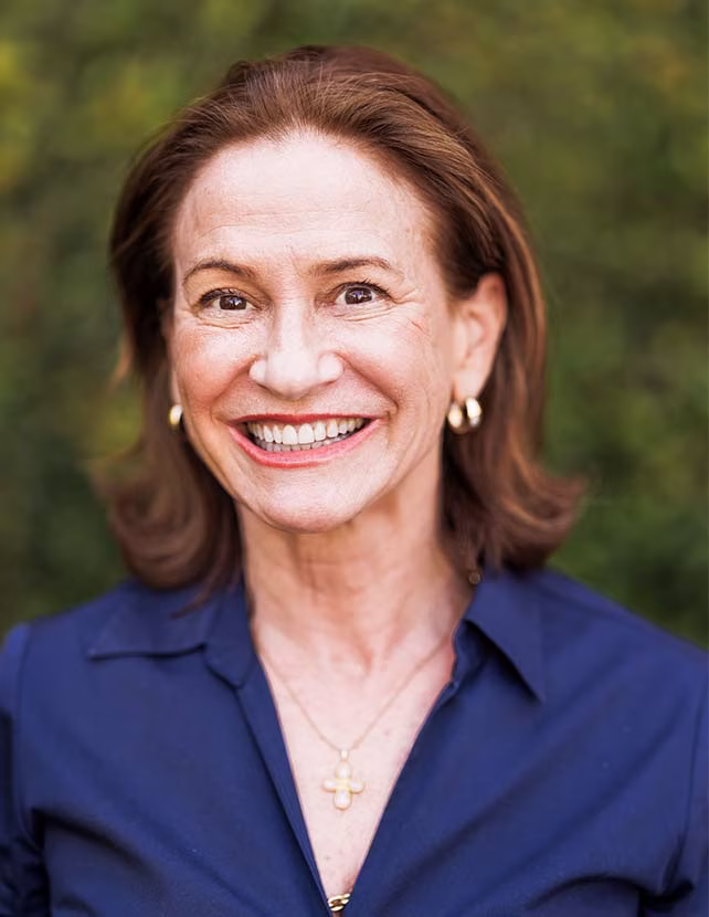A woman in a blue button down is smiling. Green leaf background.