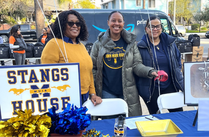 Three African American women stand at a table with a sign that says STANGS HOUSE and has festive blue and yellow decor