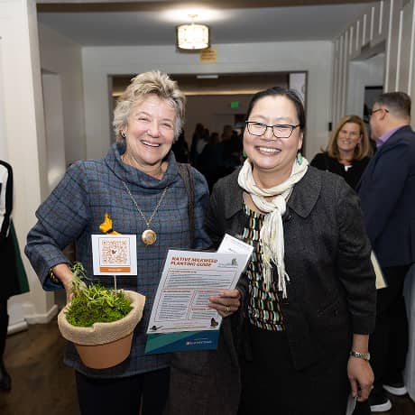 Two women smiling for a photo, one holding a plant and flyer in her hands