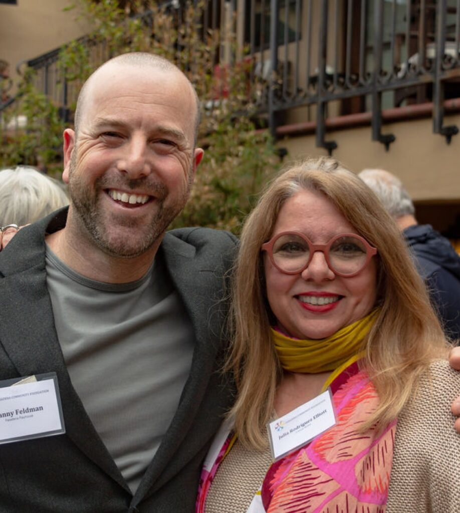 Two adults, a man and woman, stand outdoors with a balcony visible behind them.